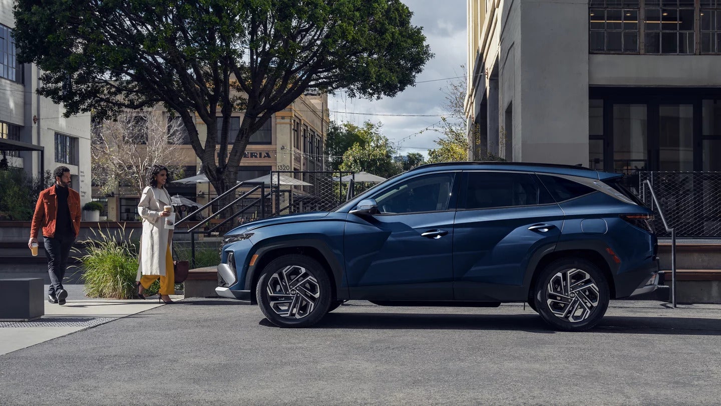 A parked, blue Hyundai Tucson in profile with a couple walking towards it